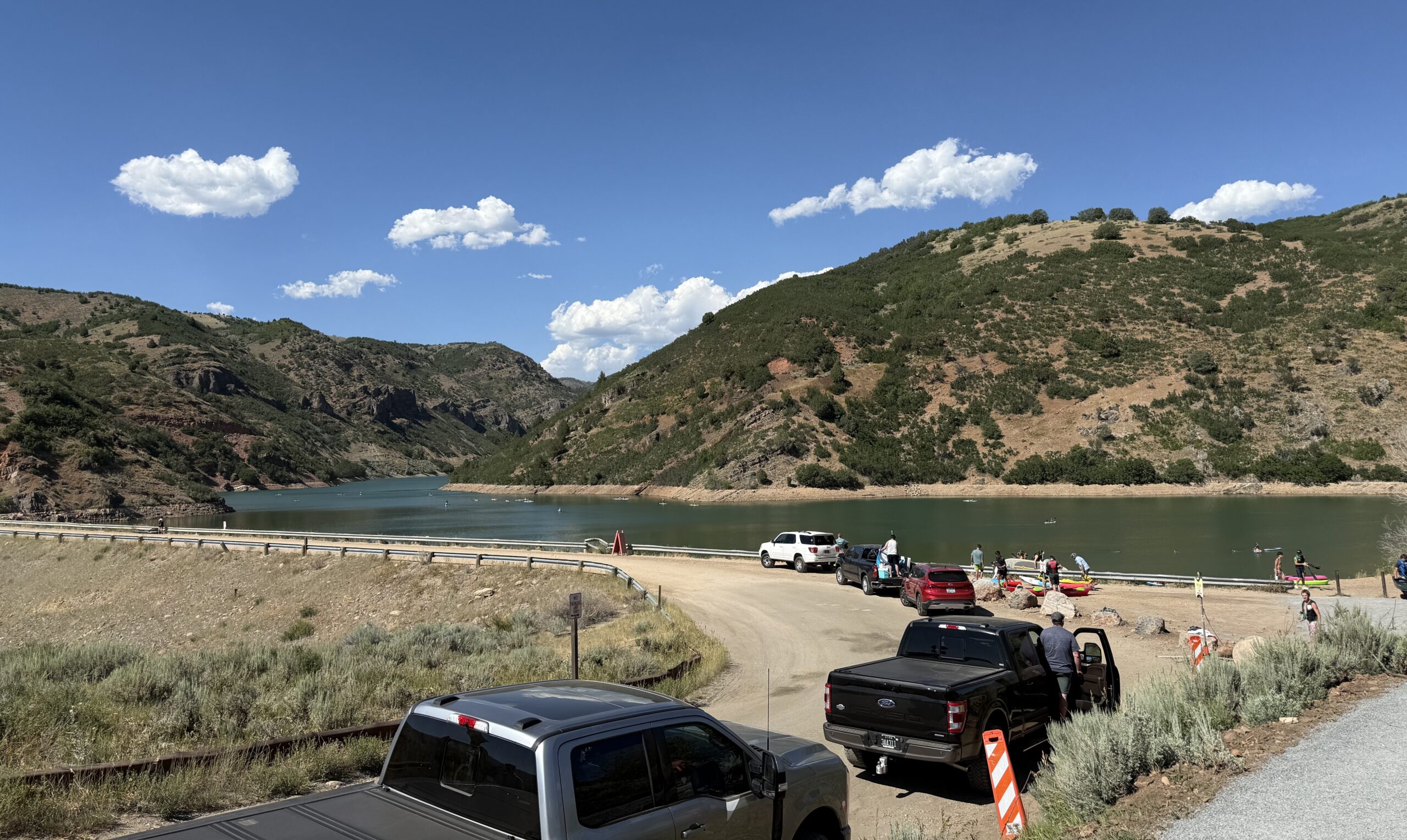 A view of Causey Dam on a busy evening. There is a line of trucks and cars along the roadside in a no parking zone. People are actively getting out of vehicles and carrying paddleboard gear to the water. The reservoir is nestled between hills covered in vegetation with a vibrant blue sky spotted with fluffy clouds.