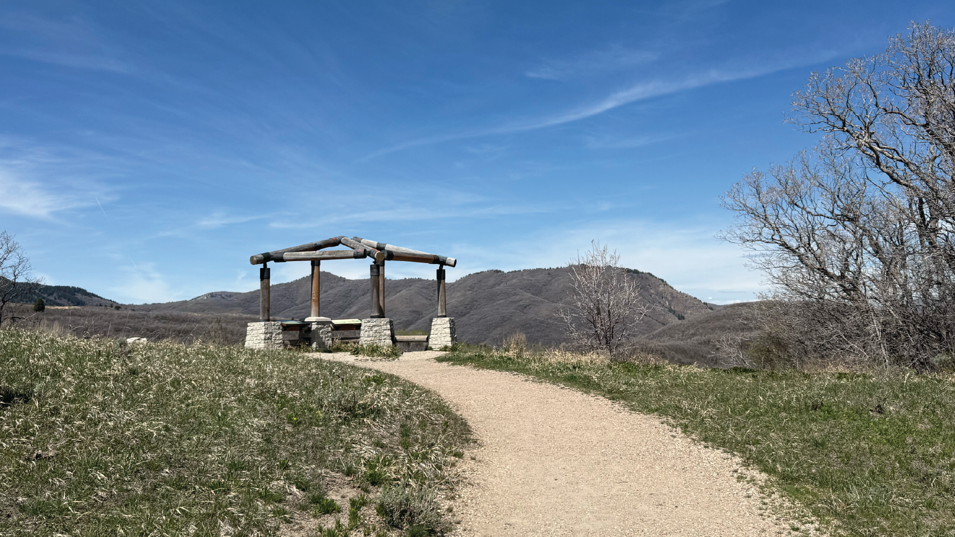 A tranquil outdoor observation point situated along a dirt path, designed to offer visitors a clear view of the surrounding mountainous landscape. The centerpiece is an open-air timber pavilion supported by stone pedestals, which provides a structured space amidst the rolling hills.