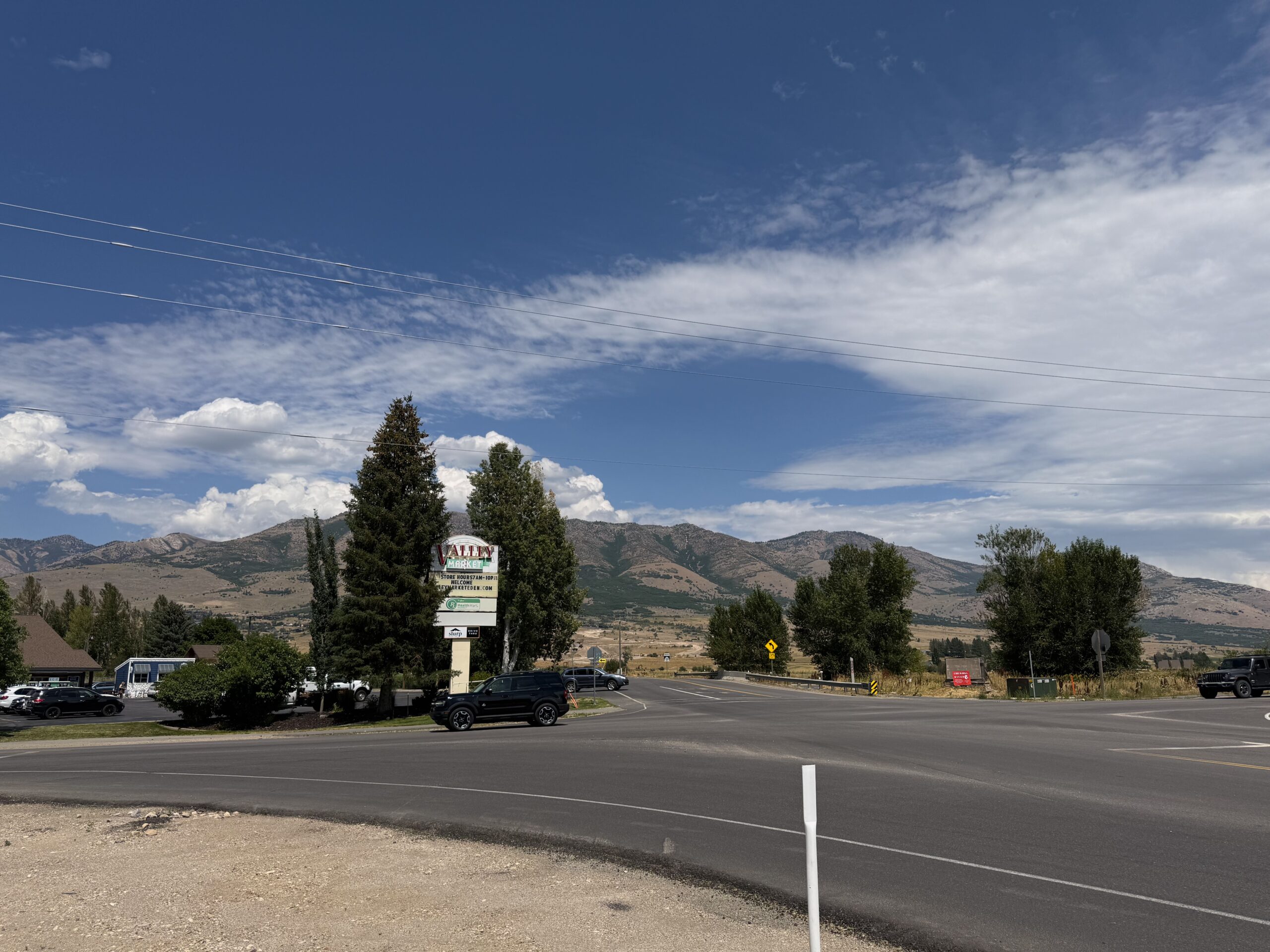 A scenic landscape photograph of a valley featuring a reservoir and sun-drenched mountains under a clear sky. In the foreground, a wide field of dry, golden-brown grass stretches toward a paved two-lane road.
