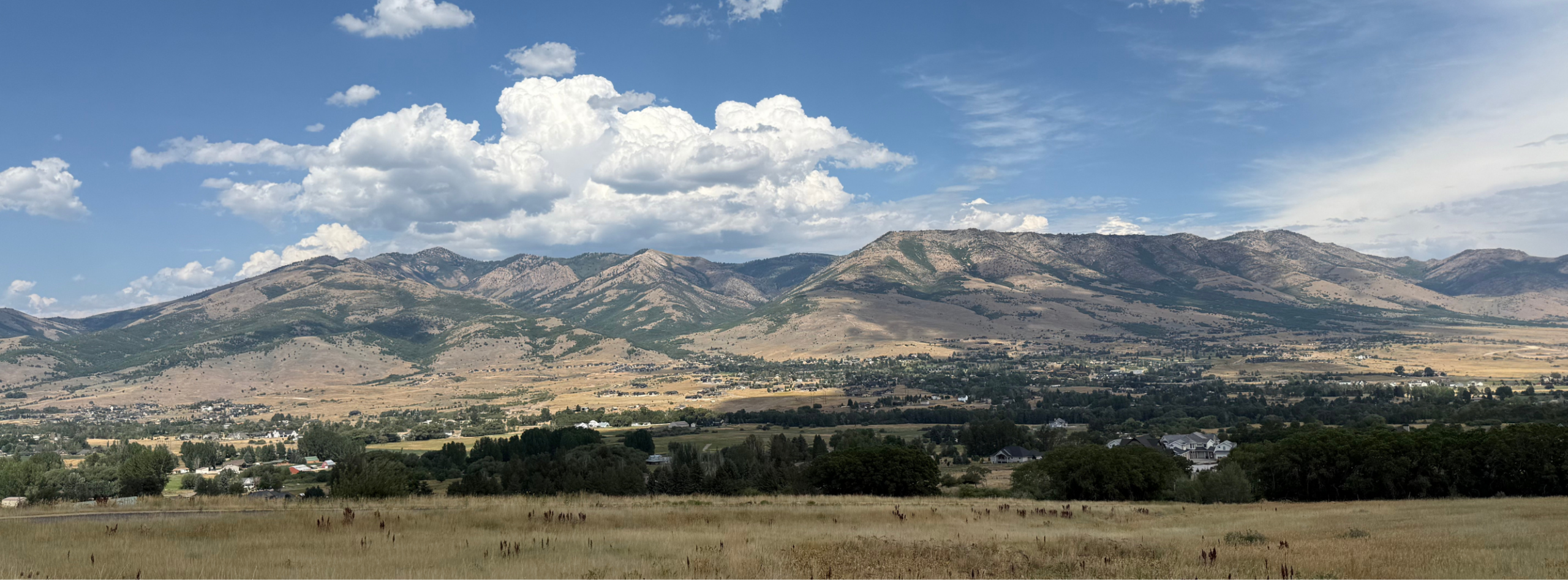 A wide-angle photograph capturing a scenic mountain landscape on a partly cloudy day. The photo has a darker, shadowed area of evergreens in the foreground framing a vast valley that recedes up the mountains. A bold mountain range is in the background