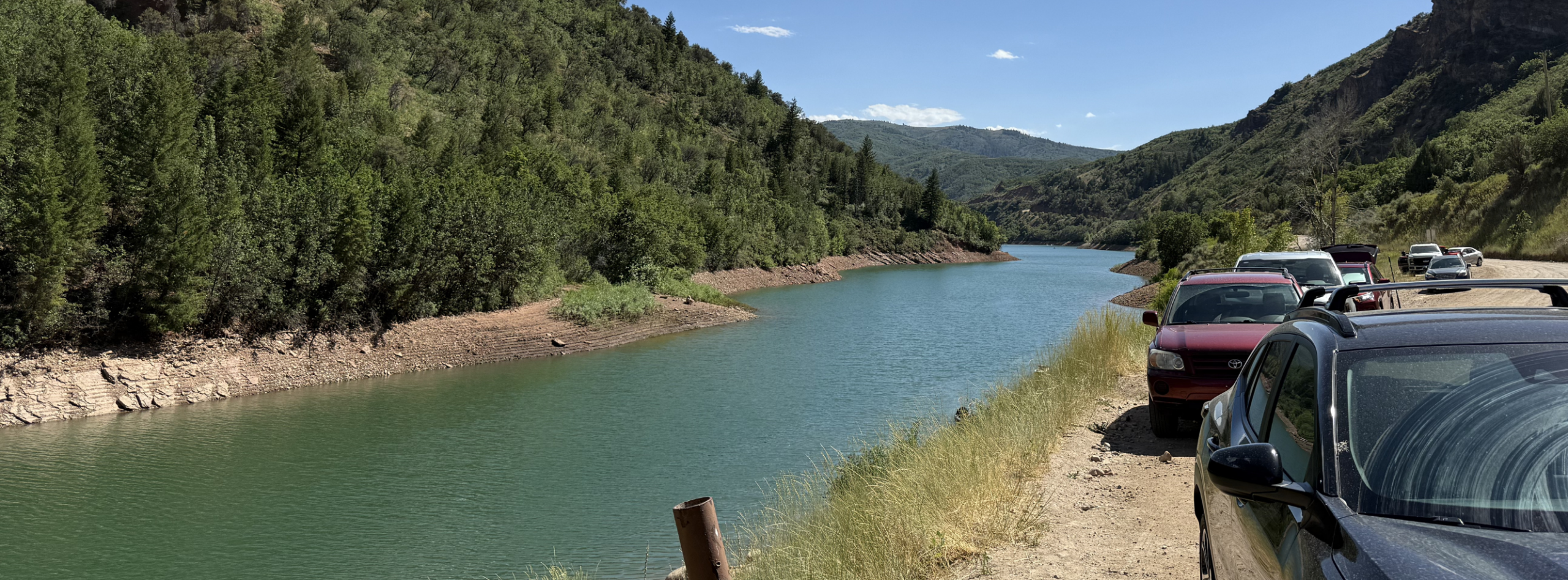 A scenic image of a narrow reservoir nestled within a steep, mountainous landscape covered in dense green vegetation. Along the right side of the frame, several parked vehicles line a dusty shoulder showing that Causey Reservoir is a popular outdoor recreation destination.