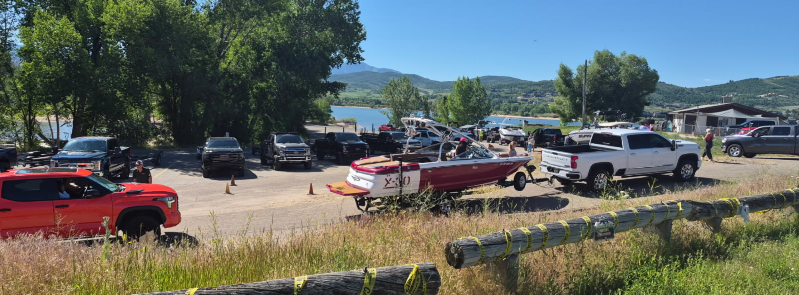 Cemetery Point at Pineview Reservoir on a busy day. The parking lot is full of trucks and boat trailers lining up for an AIS inspection prior to launching into the water. A white truck pulling a red and white speed boat followed by a red truck appear as the center focus of the image. A log fence wrapped in yellow caution tape sits in the foreground.Mountains and the lake are visible in the background.