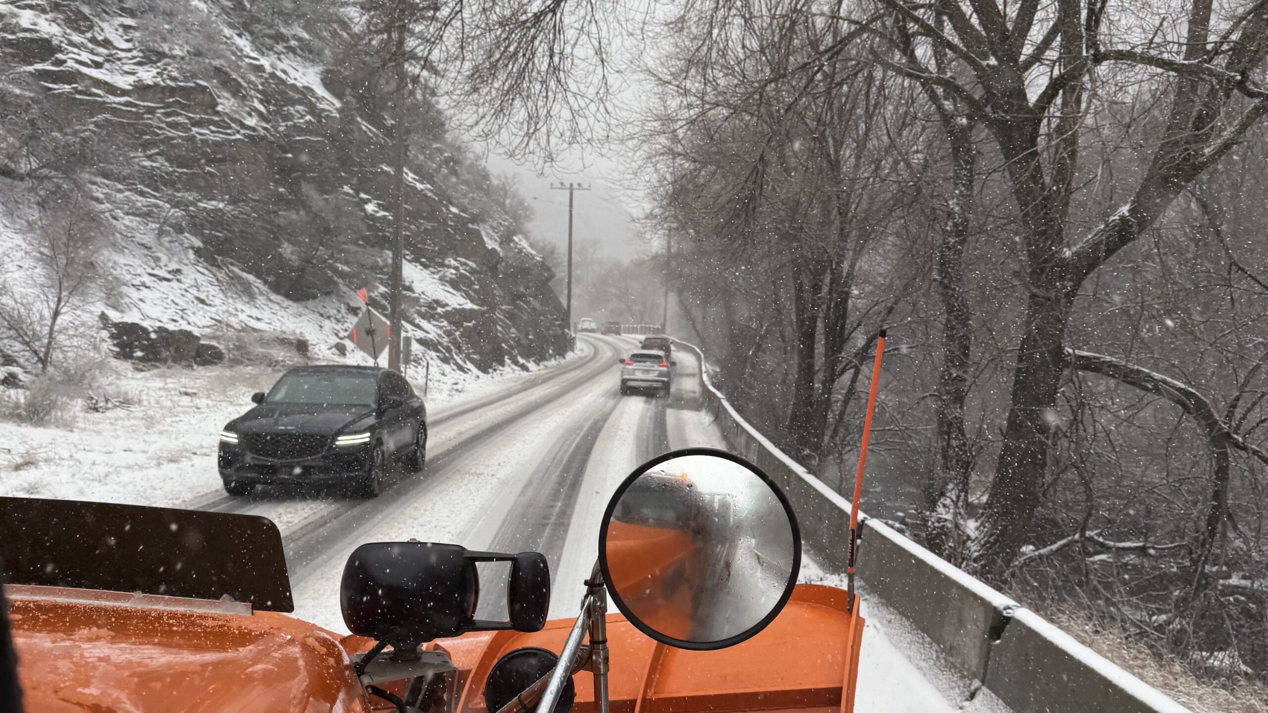 A road through a mountain canyon with traffic during a winter storm, with mountain slopes and trees in the background.